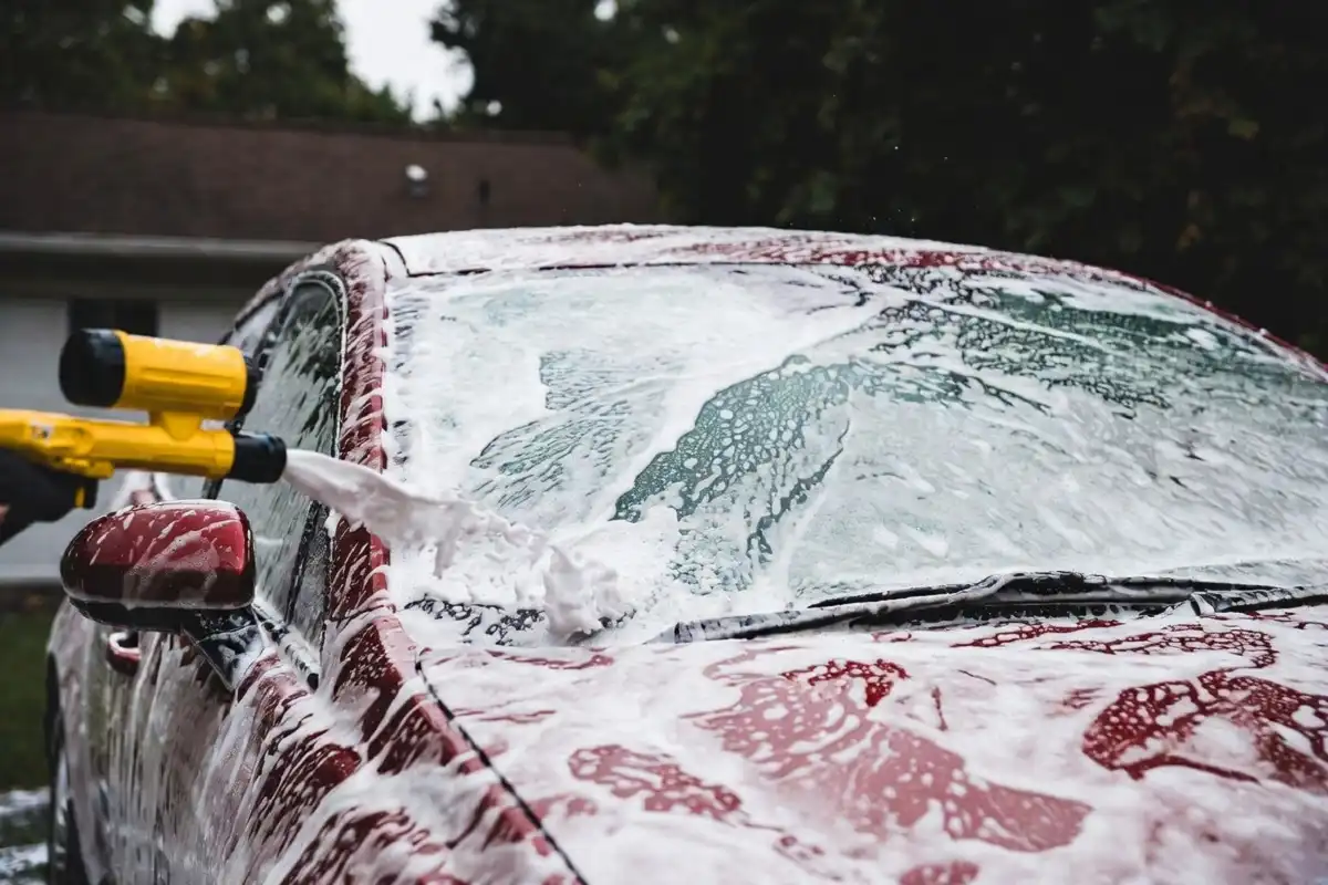Foam cannon spraying thick white soap foam on a red car as part of a home car wash machine setup