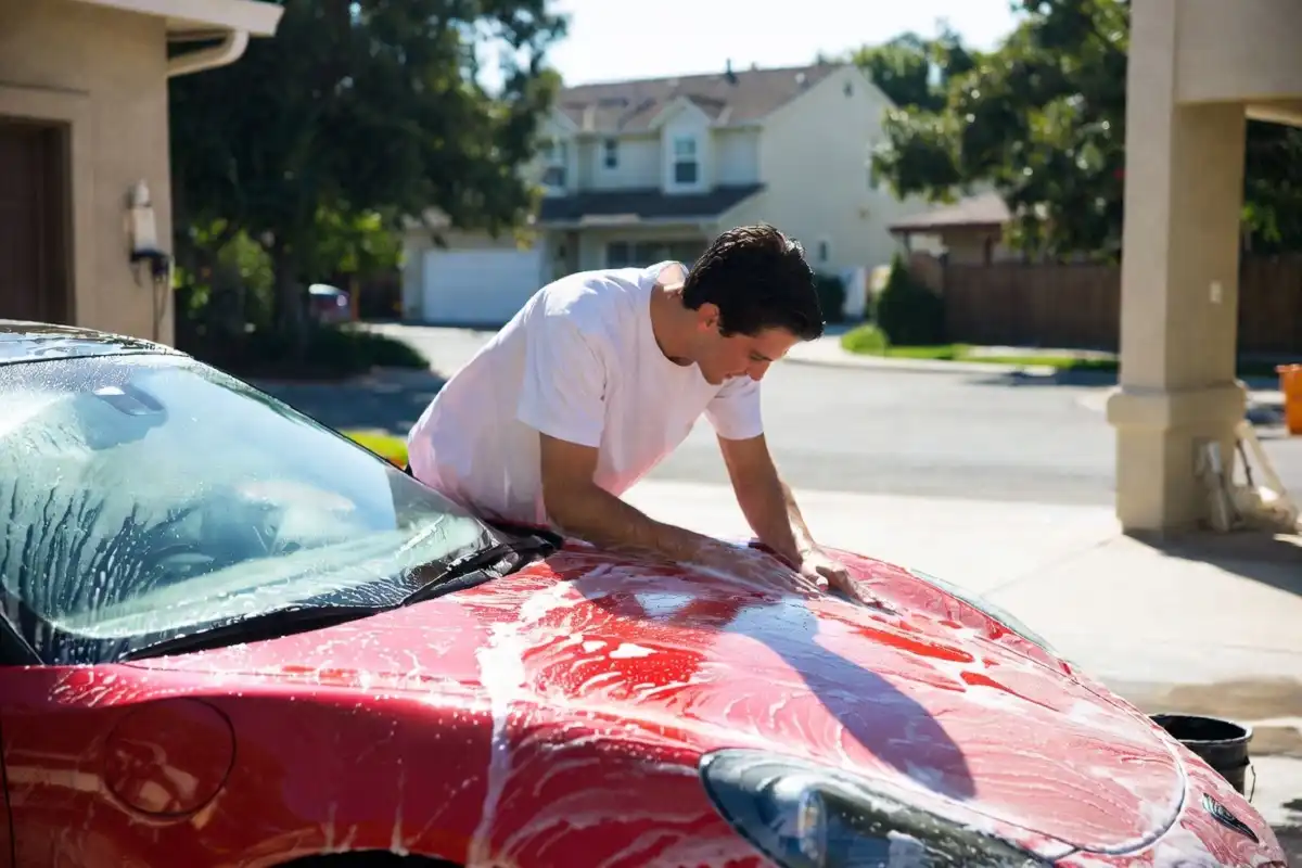 Person performing a hand car wash on a red sports car in bright sunlight