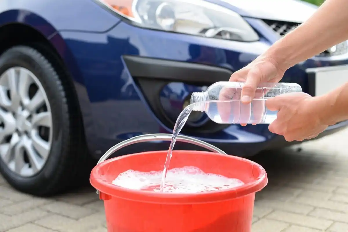 Person mixing homemade car wash soap with vinegar in a red bucket outdoors next to a car