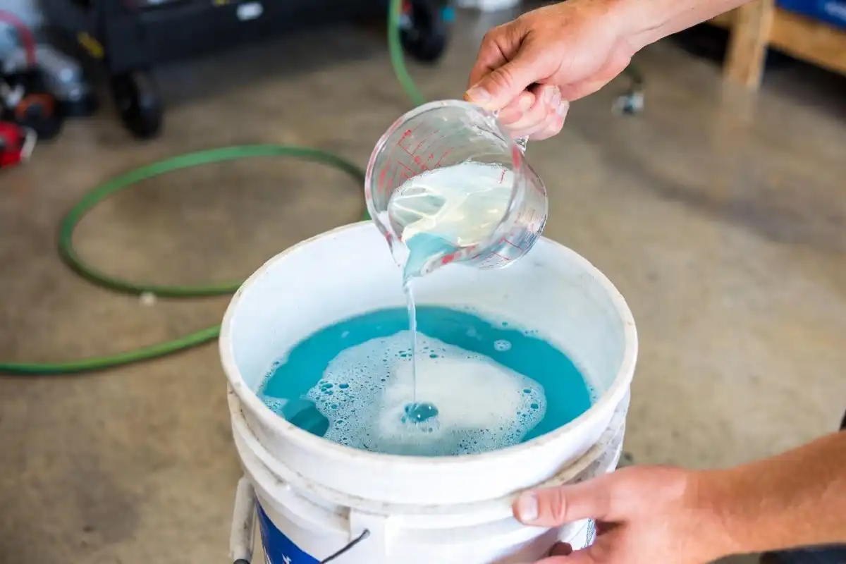 Hands pouring white vinegar into a bucket with dish soap to make homemade car wash soap