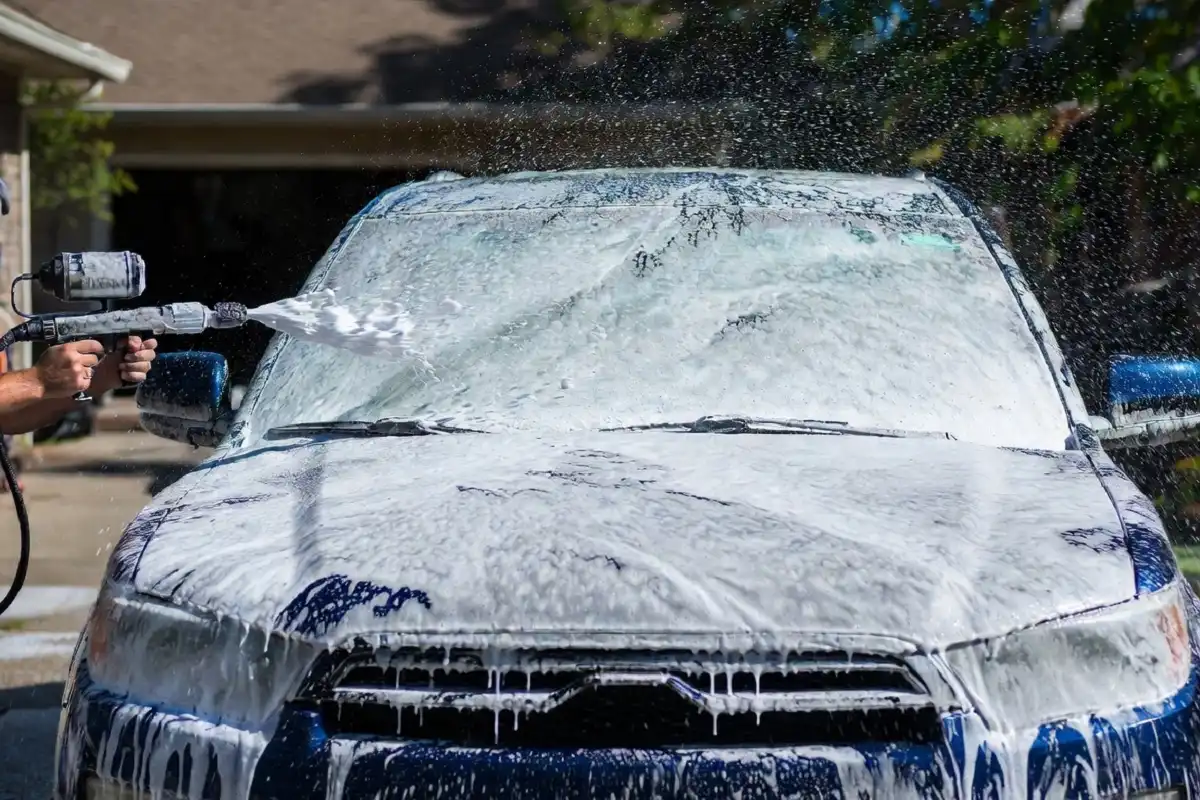 Person using a foam cannon to apply soap to a dark blue SUV during a car wash